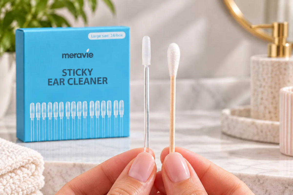 Hands holding a clear sticky ear cleaner stick and a cotton bud side by side above a box of ear cleaners in a bathroom