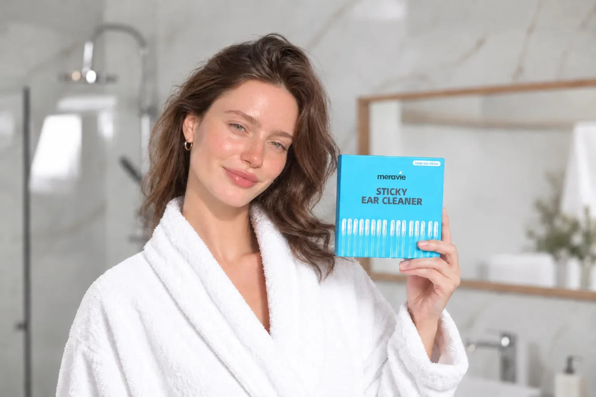 Smiling woman in a white robe holding a box of sticky ear cleaners in a bright bathroom with a glass shower behind her